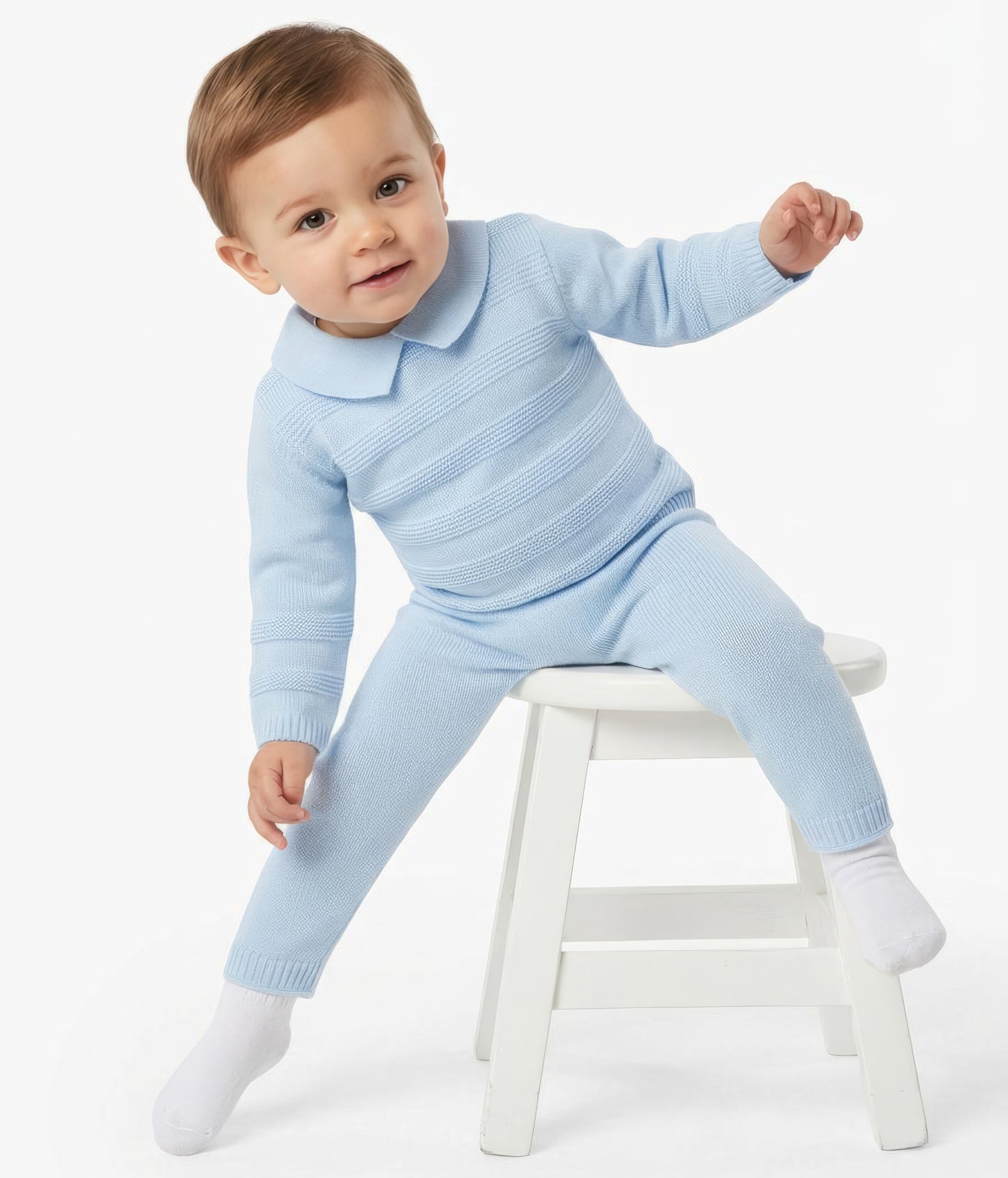 A baby wearing a light blue ribbed knit one-piece outfit with a collar and matching pants, sitting on a small white stool against a plain white background.