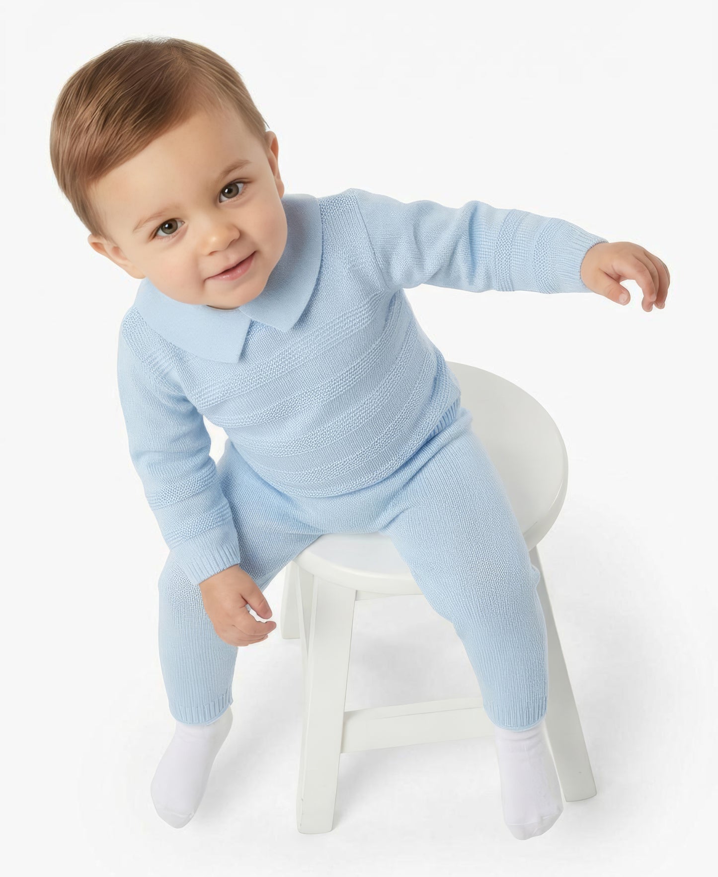 A baby wearing a light blue ribbed knit one-piece sweater and matching pants, sitting on a small white stool against a plain white studio background.
