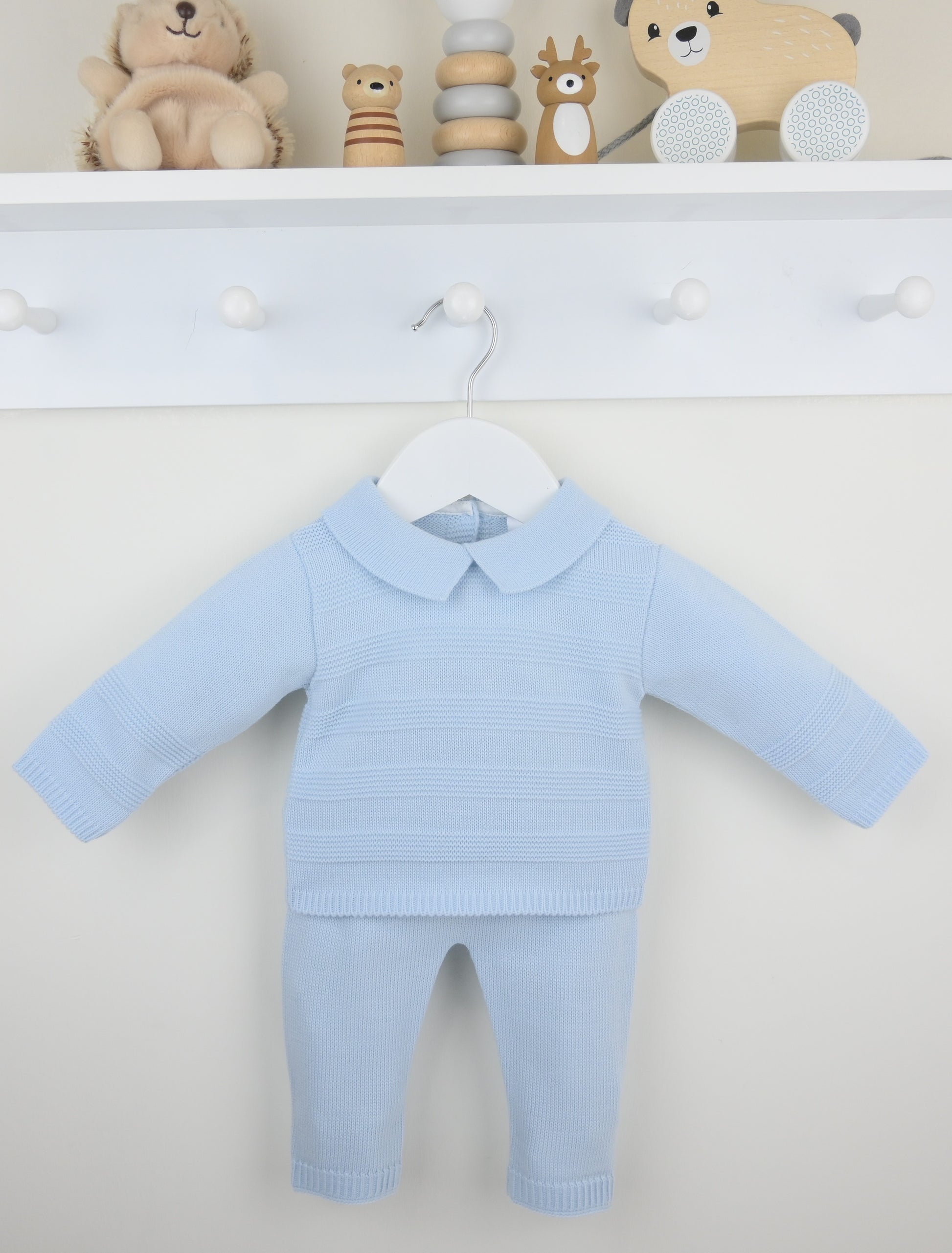 A baby two-piece knit outfit in light blue, featuring a long-sleeve top with a Peter Pan collar and matching pants, hanging on a white hanger against a white wall with toy decorations on a shelf above.
